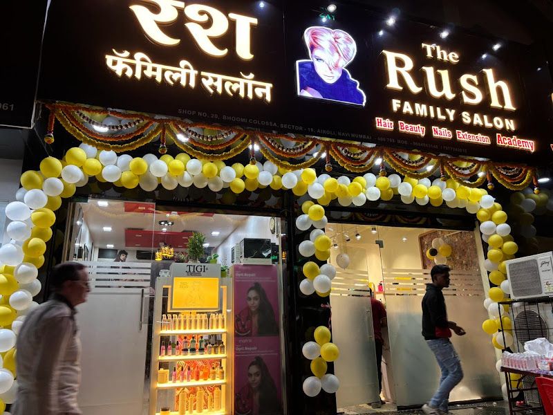 Evening exterior of the salon — illuminated 'The Rush Family Salon' signage (English + Marathi 'रश फॅमिली सलॉन') with the gold crown-R logo, glass frontage and a yellow-and-white balloon arch from an event/opening. — The Rush Family Salon, Airoli, Navi Mumbai