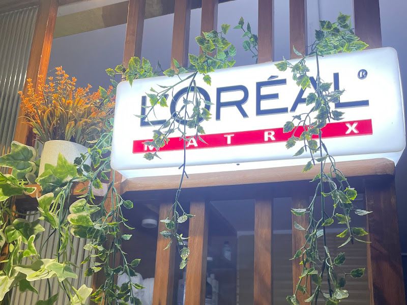 Interior brand corner — illuminated L'Oréal / Matrix sign framed by trailing money-plant greenery against a slatted wood wall. — The Rush Family Salon, Airoli, Navi Mumbai