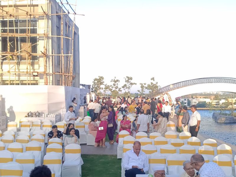 Large outdoor / waterfront event — rows of white chairs with gold sashes on a lawn, guests seated, a bridge in the background — Arya Events Planner, Kharghar, Navi Mumbai