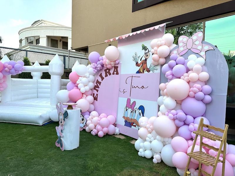 Minnie / Daisy theme outdoor 2nd birthday ('Emira is Two') — pastel balloon wall with character cutouts beside a white bouncy castle — Arya Events Planner, Kharghar, Navi Mumbai