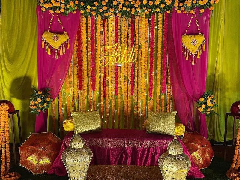 Haldi stage — magenta and marigold floral backdrop with a gold 'Haldi' sign, low seating, decorative umbrellas and brass urli bowls — Arya Events Planner, Kharghar, Navi Mumbai