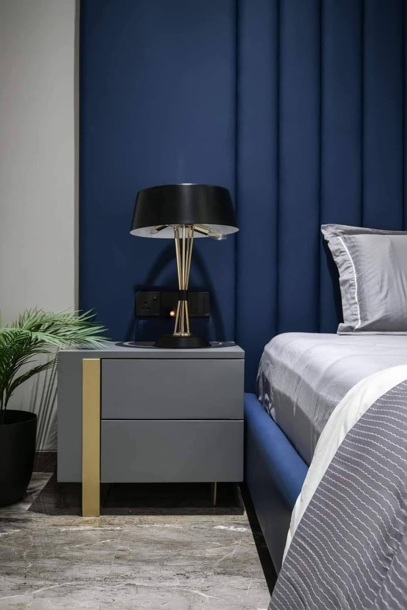 Bedroom with blue upholstered bed, bedside table, lamp, plant and gray bedding